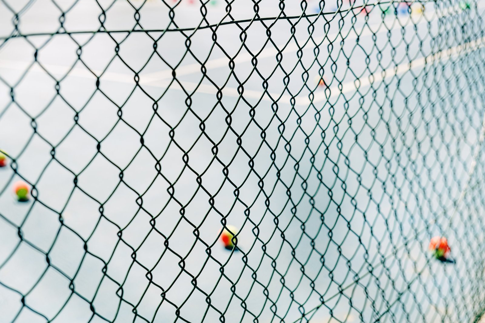 Background of a metal net fence in the foreground on a tennis court with defocused balls on the ground.